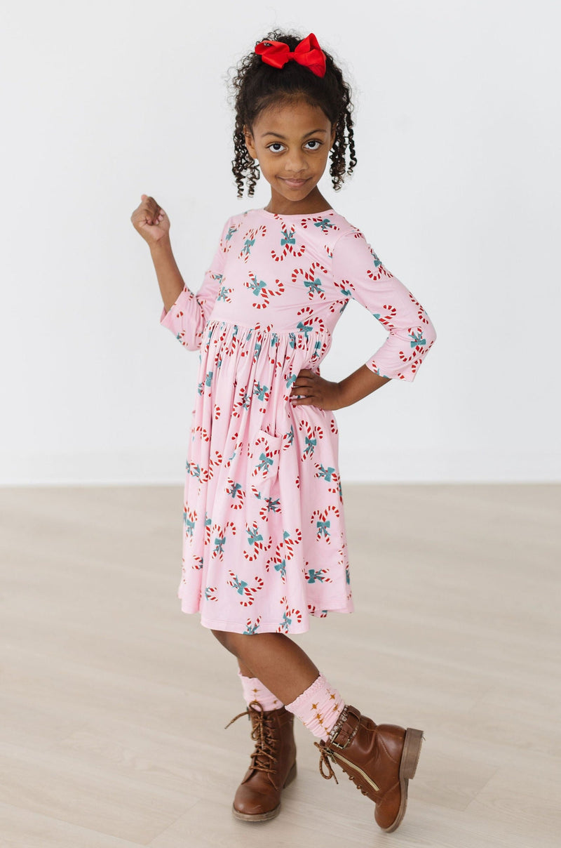 Child confidently posing in the Candy Cane Cutie 3/4 Sleeve Pocket Twirl Dress by Mila & Rose, complemented by stylish brown boots, embracing holiday spirit.