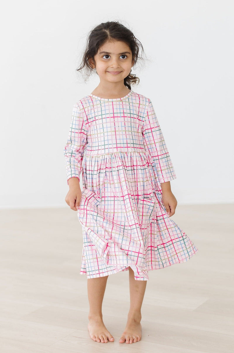 A joyful girl in a Pink Plaid 3/4 Sleeve Pocket Twirl Dress, standing barefoot and sporting a playful smile as she twirls in her stylish outfit.