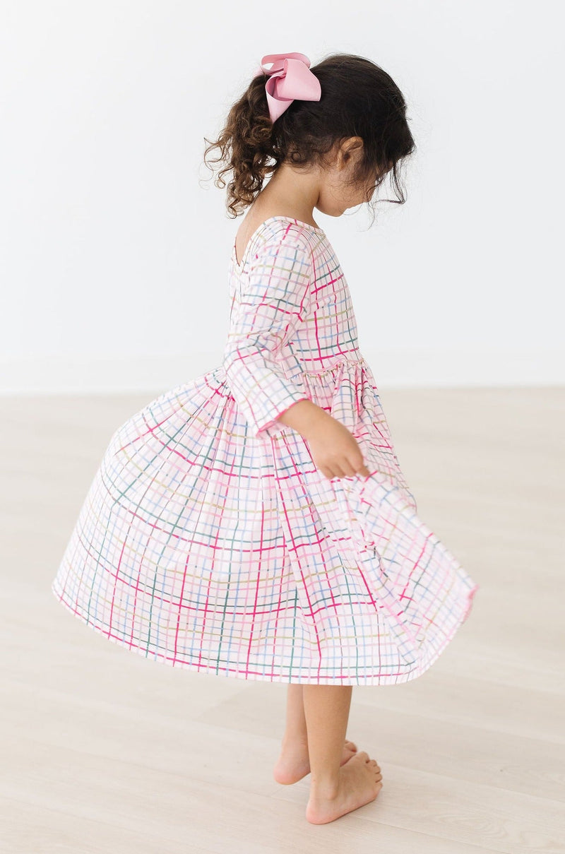 A little girl spins gracefully in her Pink Plaid 3/4 Sleeve Pocket Twirl Dress, beautifully highlighting its twirly design paired with a lovely pink bow in her curly hair.