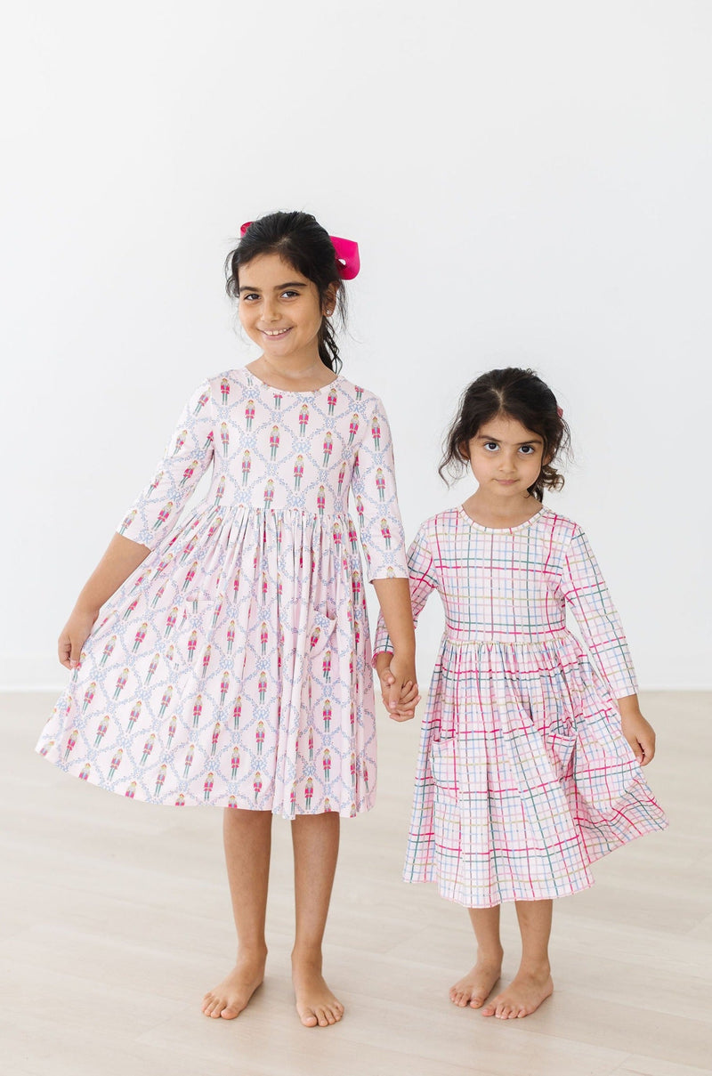 Two sisters hold hands, both in stylish dresses; one in a Pink Plaid 3/4 Sleeve Pocket Twirl Dress, radiating joy and sibling love in a stylish photoshoot.
