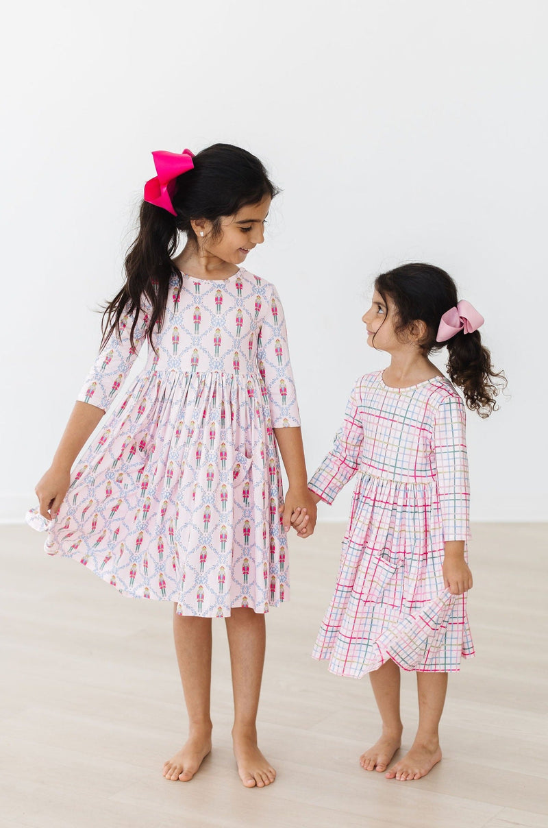 Two cheerful girls holding hands, featuring one in the Pink Nutcracker 3/4 Sleeve Pocket Twirl Dress and the other in a matching checkered twirl dress, perfect for festive fun!