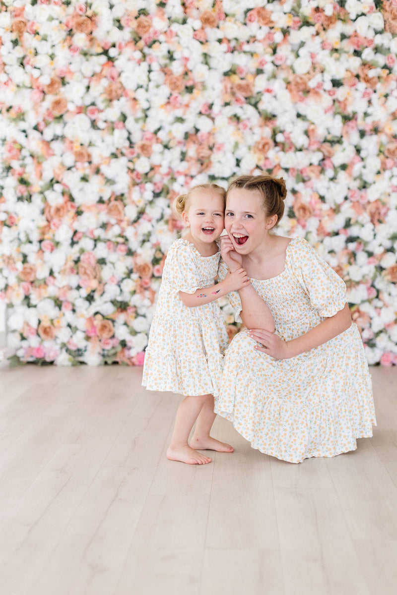 Two girls joyfully posing together wearing the Marvelous Marigolds Smocked Ruffle Dress by Mila & Rose, surrounded by a floral backdrop.