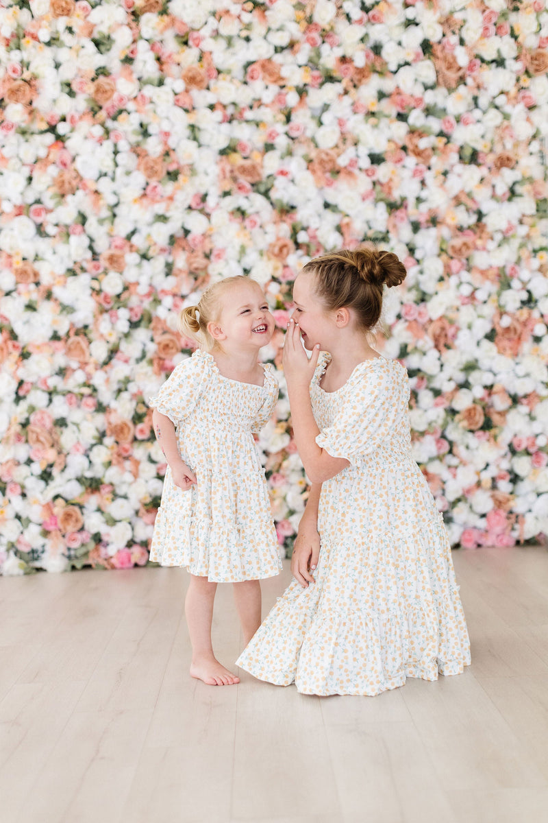 Sisters sharing a secret while wearing the Marvelous Marigolds Smocked Ruffle Dress, surrounded by a beautiful floral backdrop, emphasizing their playful bond.