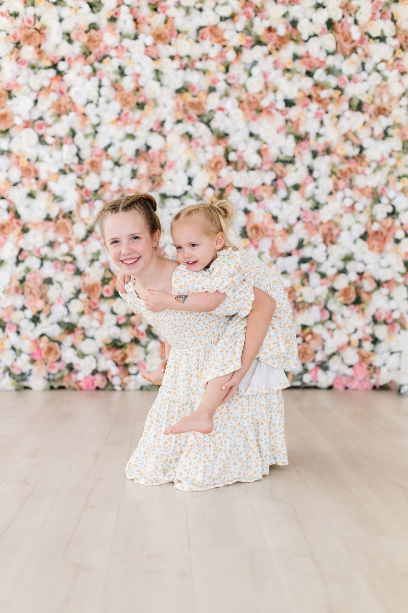 A girl being playfully carried by another girl, both in the Marvelous Marigolds Smocked Ruffle Dress by Mila & Rose, with a beautiful floral wall in the background.