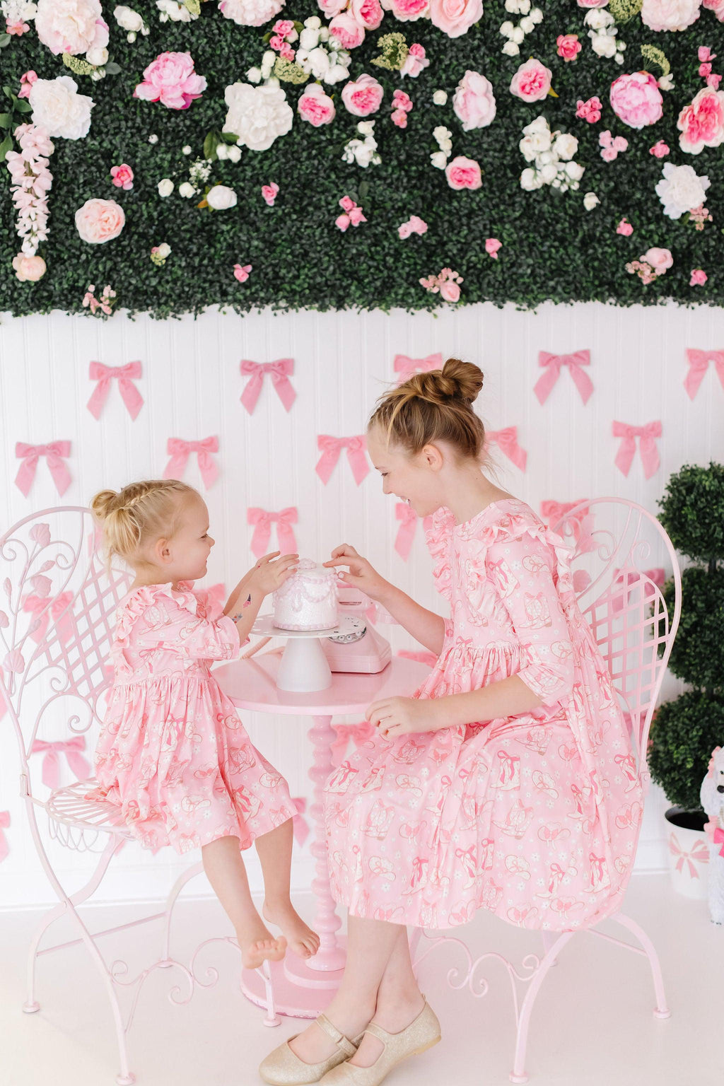 Charming scene featuring two girls wearing the Howdy 3/4 Sleeve Ruffle Twirl Dress by Mila & Rose®, joyfully sharing a tea party at a decorated table with pink bows and flowers in the background.