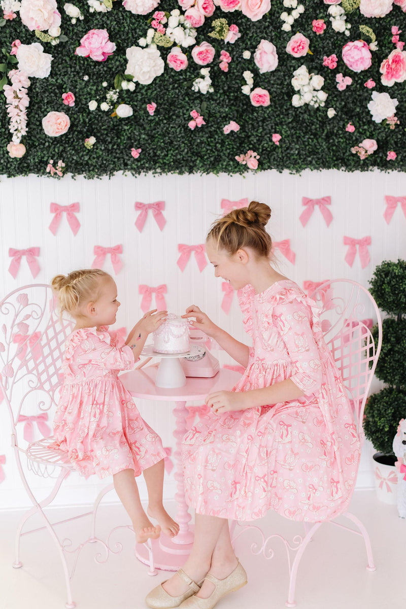 Charming scene featuring two girls wearing the Howdy 3/4 Sleeve Ruffle Twirl Dress by Mila & Rose®, joyfully sharing a tea party at a decorated table with pink bows and flowers in the background.