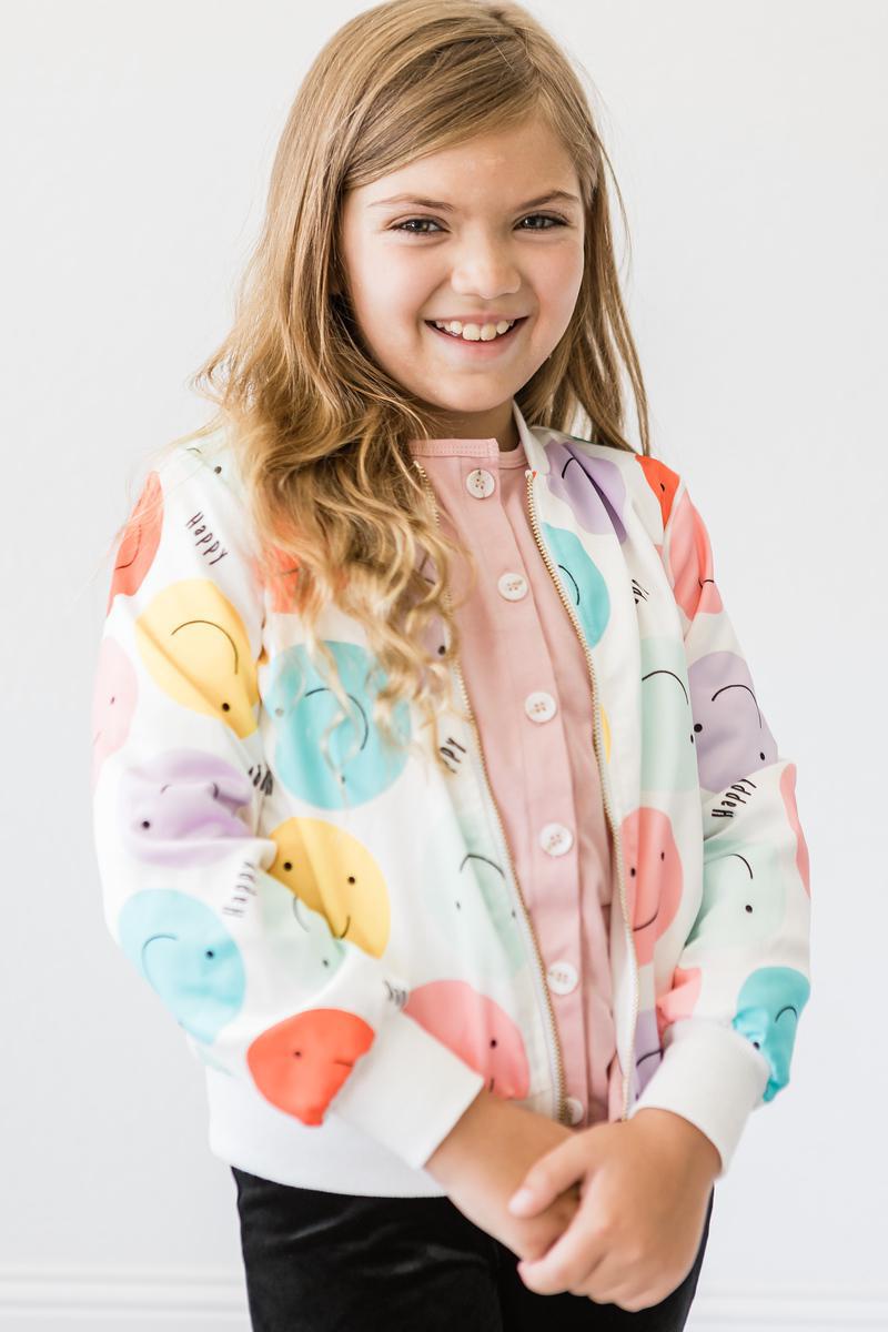 Young girl beaming in the 'Happy Days' Satin Jacket for Girls, featuring a charming design of smiley faces, perfectly paired with a pink shirt.
