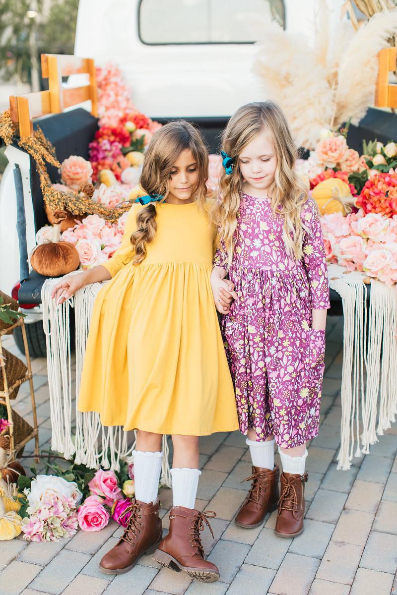 Two girls wearing the Mustard 3/4 Sleeve Pocket Twirl Dress by Mila & Rose® and a floral patterned dress, surrounded by decorative flowers and rustic elements.