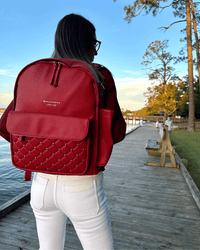Crimson vegan leather diaper bag with quilted front pocket and gold studs worn on a waterfront boardwalk.