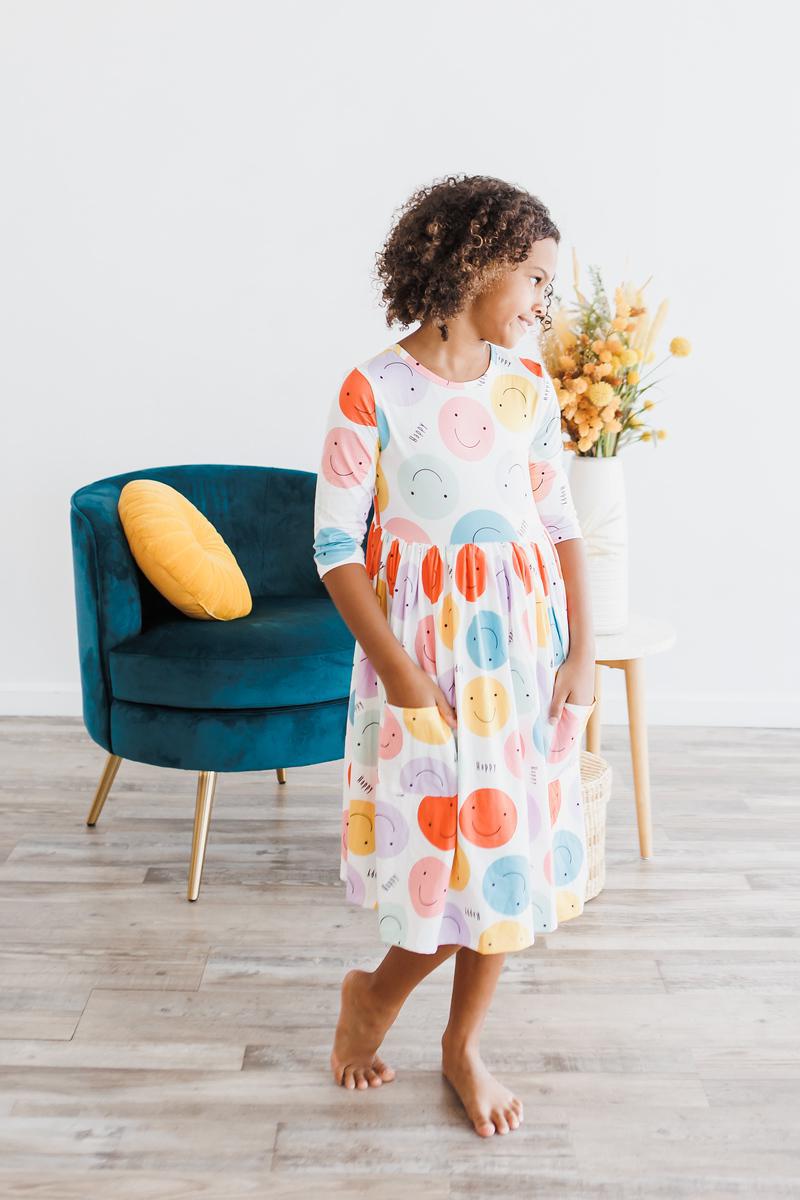 A girl posing in the 'Happy Days' 3/4 Pocket Fun Twirl Dress for Girls, decorated with vibrant smiley patterns, perfect for school or play.