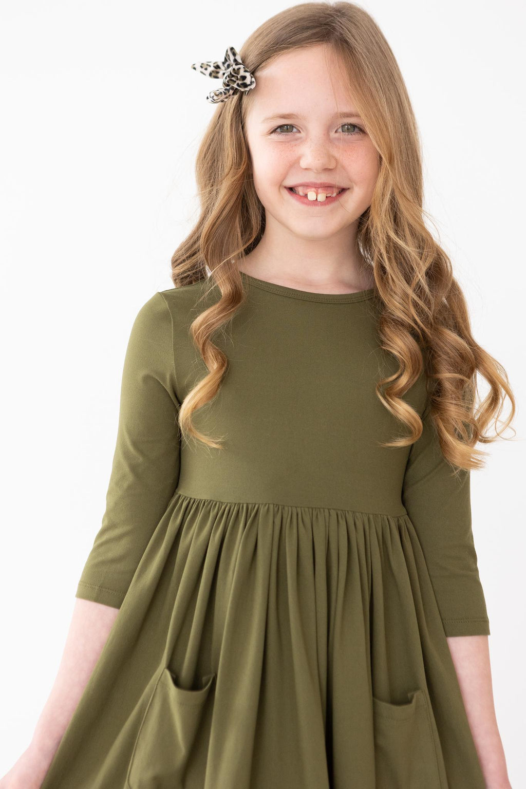 Olive 3/4 Sleeve Pocket Twirl Dress modeled by a smiling girl with wavy hair, showcasing its soft fabric and playful twirl-worthy design.