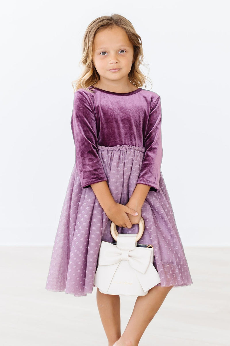 A girl standing confidently in an Amethyst Velvet Tutu Dress, featuring 3/4 sleeves and a beautiful tulle skirt, holding a chic white handbag.