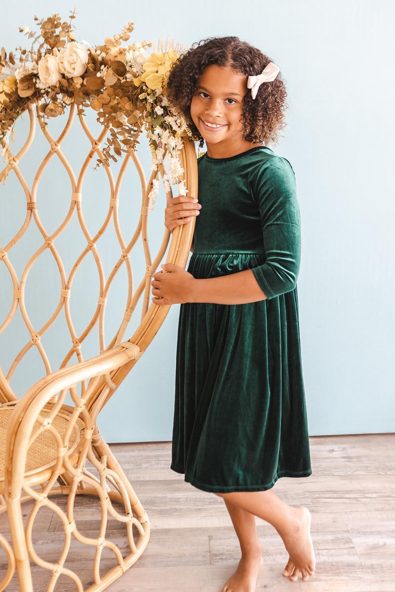 Little girl smiling in a Hunter Green Velvet Twirl Dress by Mila & Rose, standing beside a decorative chair adorned with flowers.
