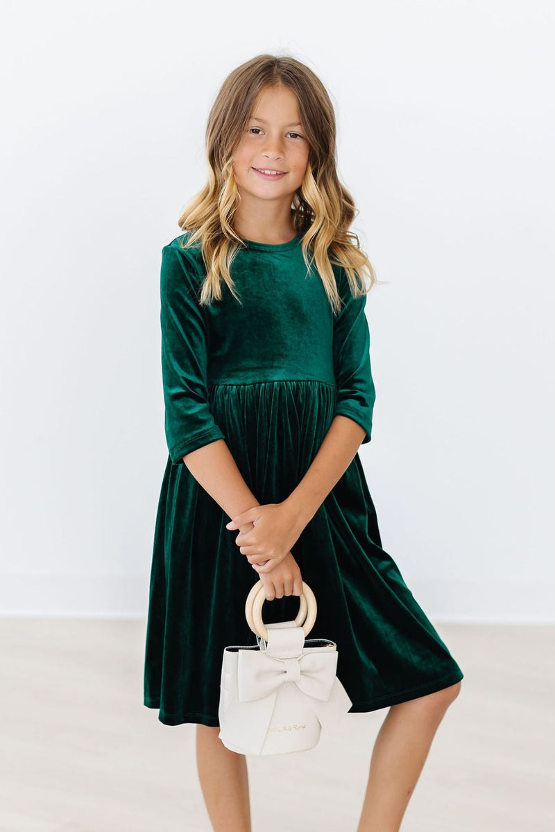 Confident young girl in a Hunter Green Velvet Twirl Dress by Mila & Rose, posing with a stylish handbag, showcasing elegance and fun.