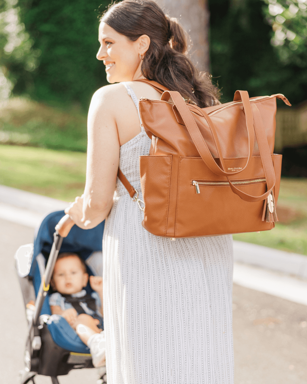 Smiling woman in white dress wearing cognac diaper bag as backpack while pushing baby stroller outdoors