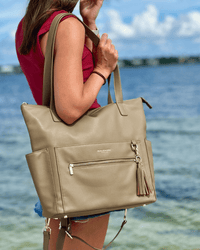 Woman in red top carrying taupe tote bag by handle against blurred waterfront background