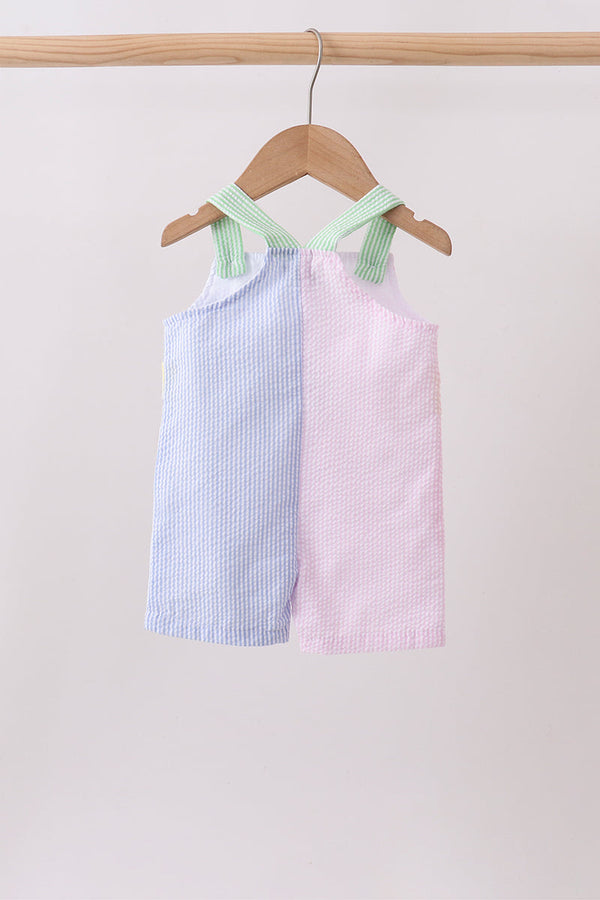 Two children's dresses, one blue and one pink, with green straps, hanging on a wooden hanger against a light background.