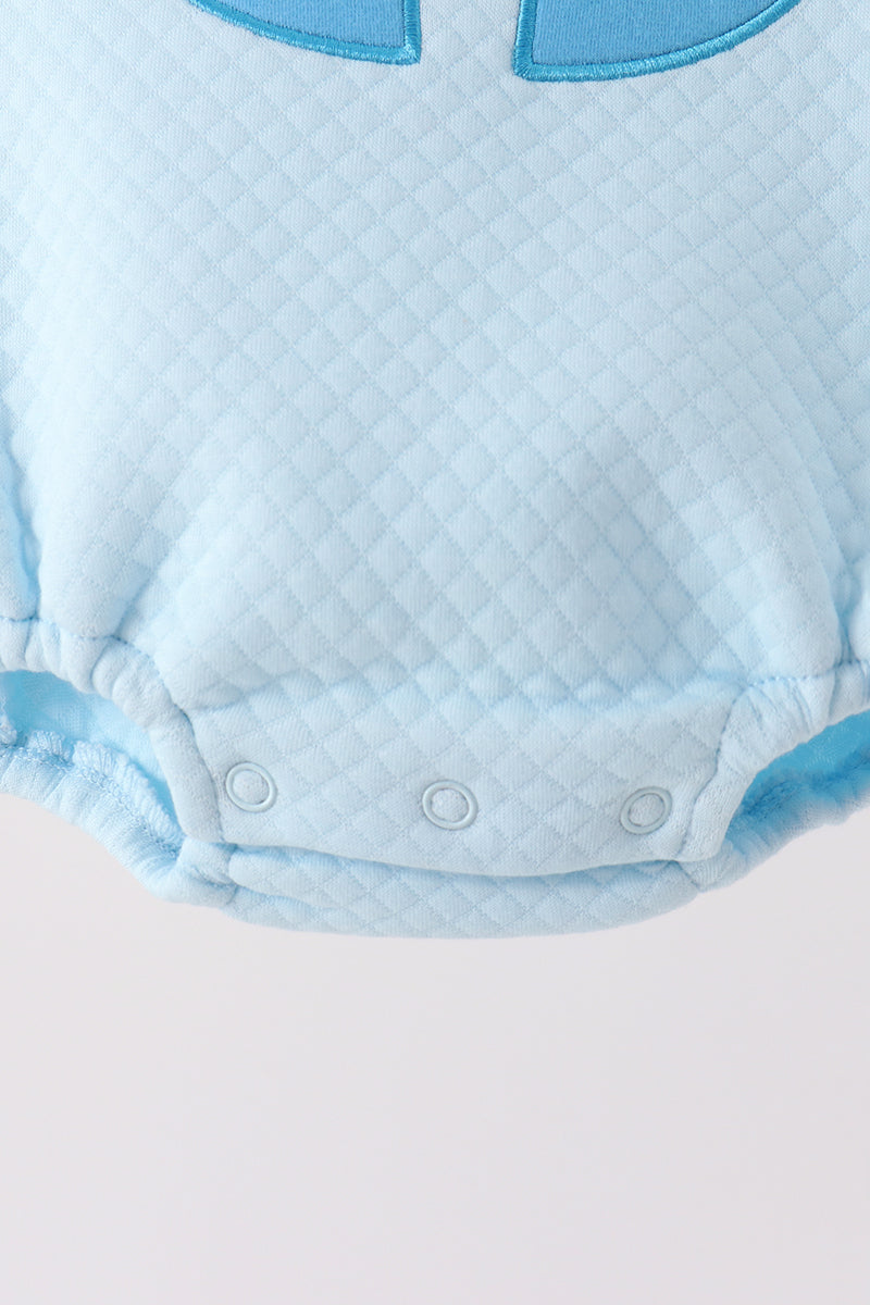 Light blue quilted baby bubble onesie featuring three snap closures at the crotch and a decorative blue appliqué trim at the neckline.