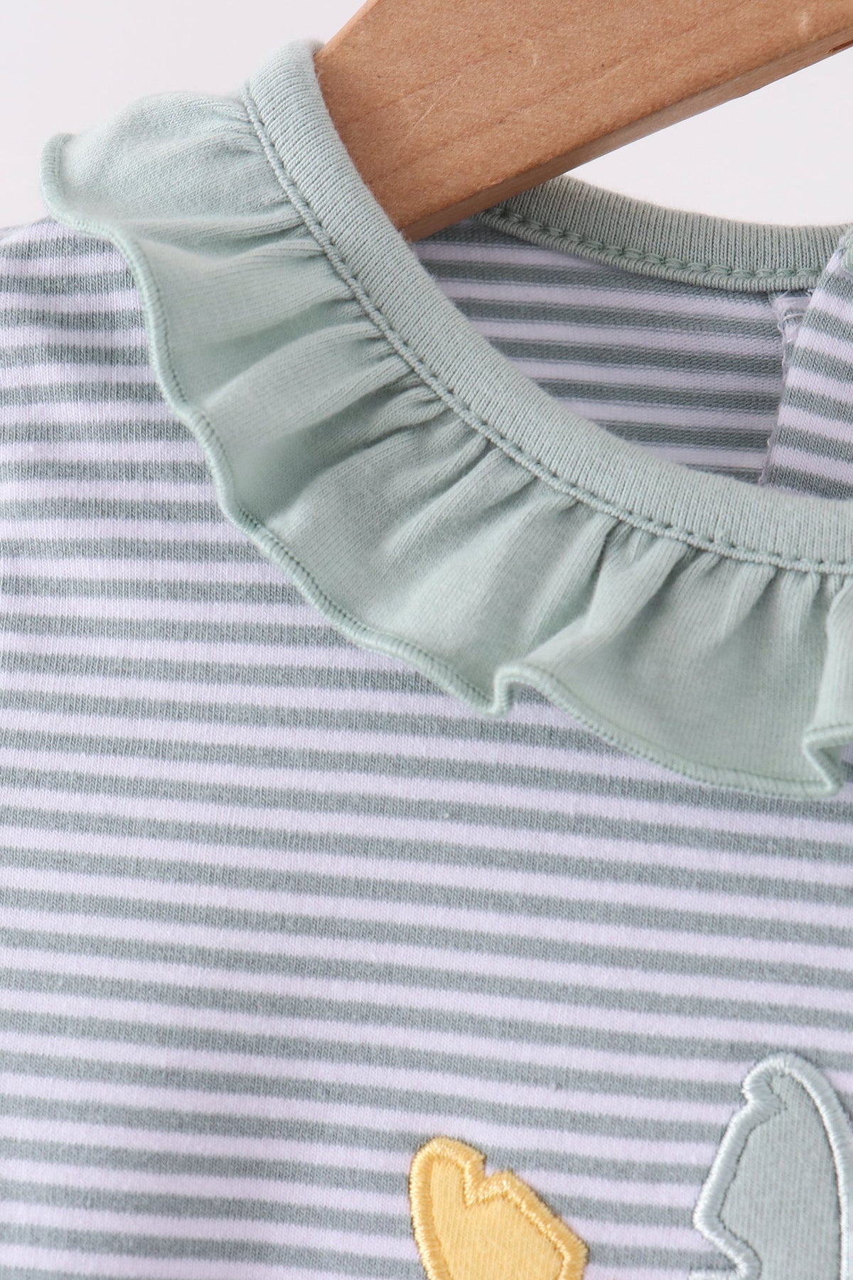 Close-up of a baby or toddler striped top on a wooden hanger, featuring a wide ruffled collar in a pale sage green and fine gray horizontal stripes.
