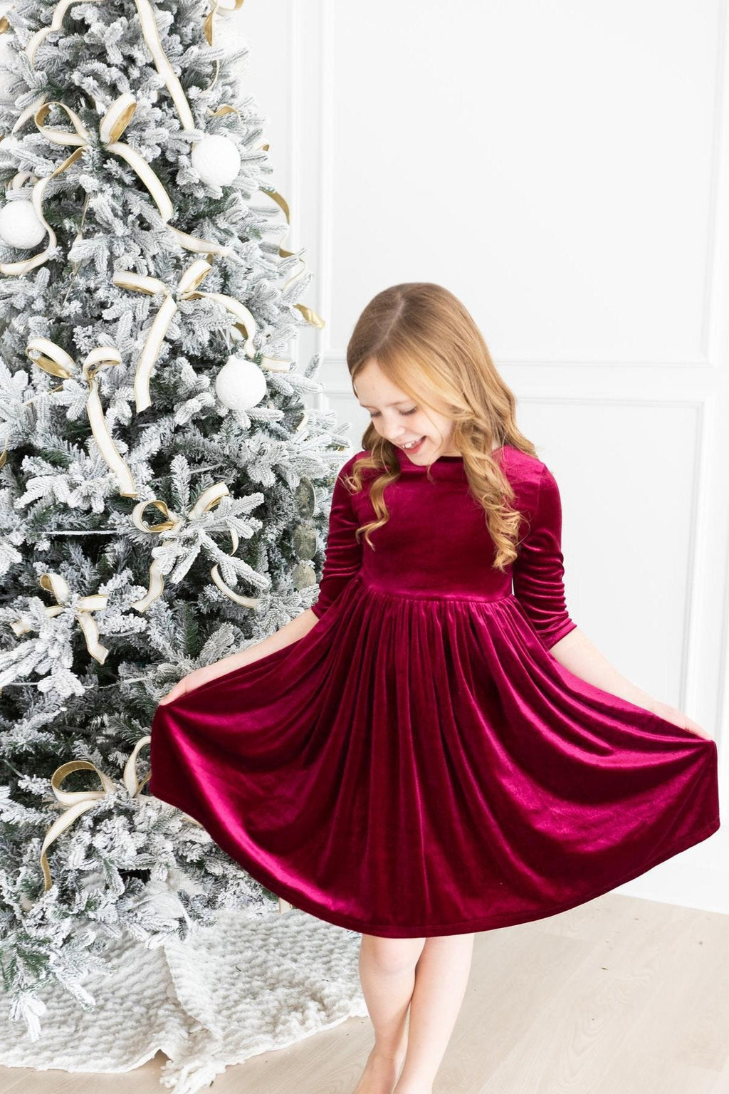 A young girl joyfully twirling in a Cranberry Velvet Twirl Dress from Mila & Rose® next to a frosted Christmas tree, capturing the spirit of holiday cheer.