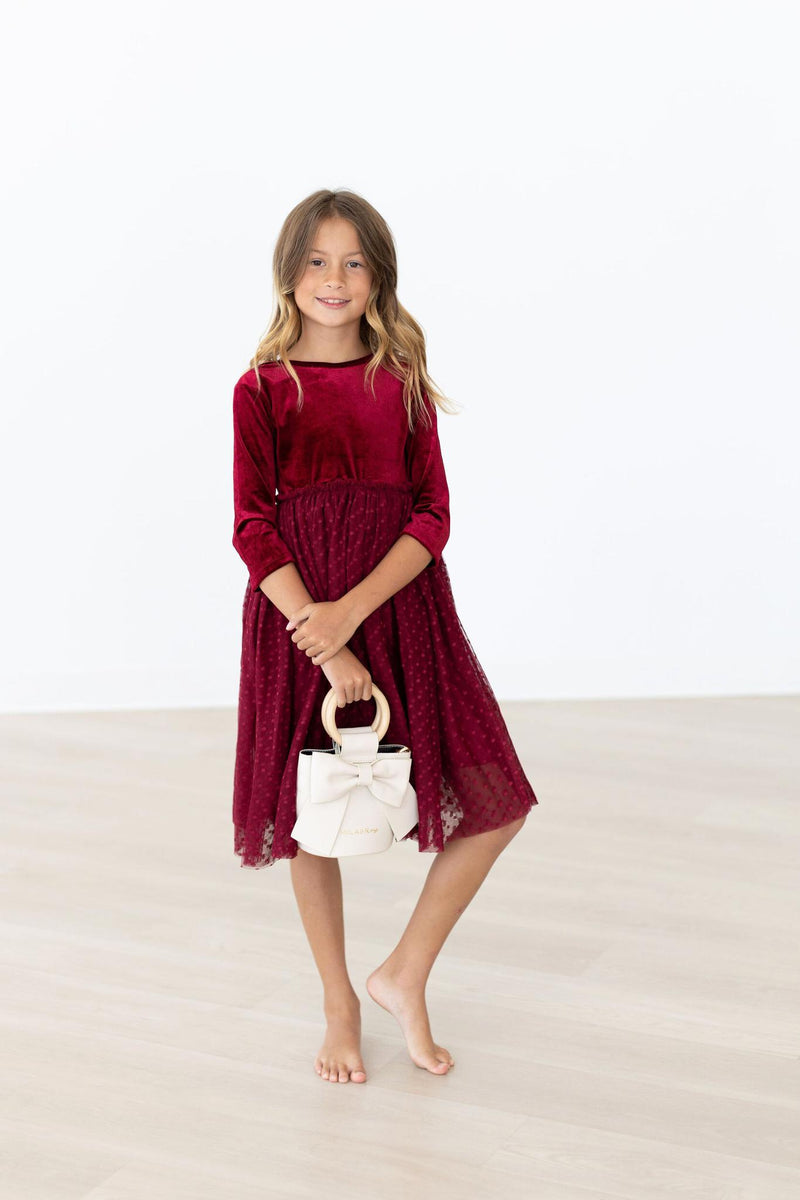 A girl posing confidently with a woven handbag, wearing a Cranberry Velvet Tutu Dress, highlighting its stylish and comfortable fit.