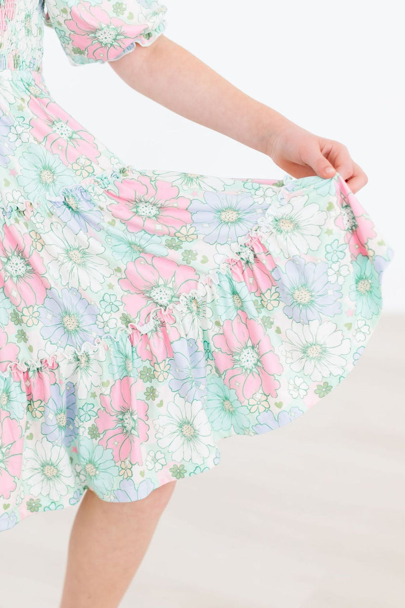 Girl holding the hem of her Minty Meadow Smocked Ruffle Dress, emphasizing the vibrant floral print and playful twirl.