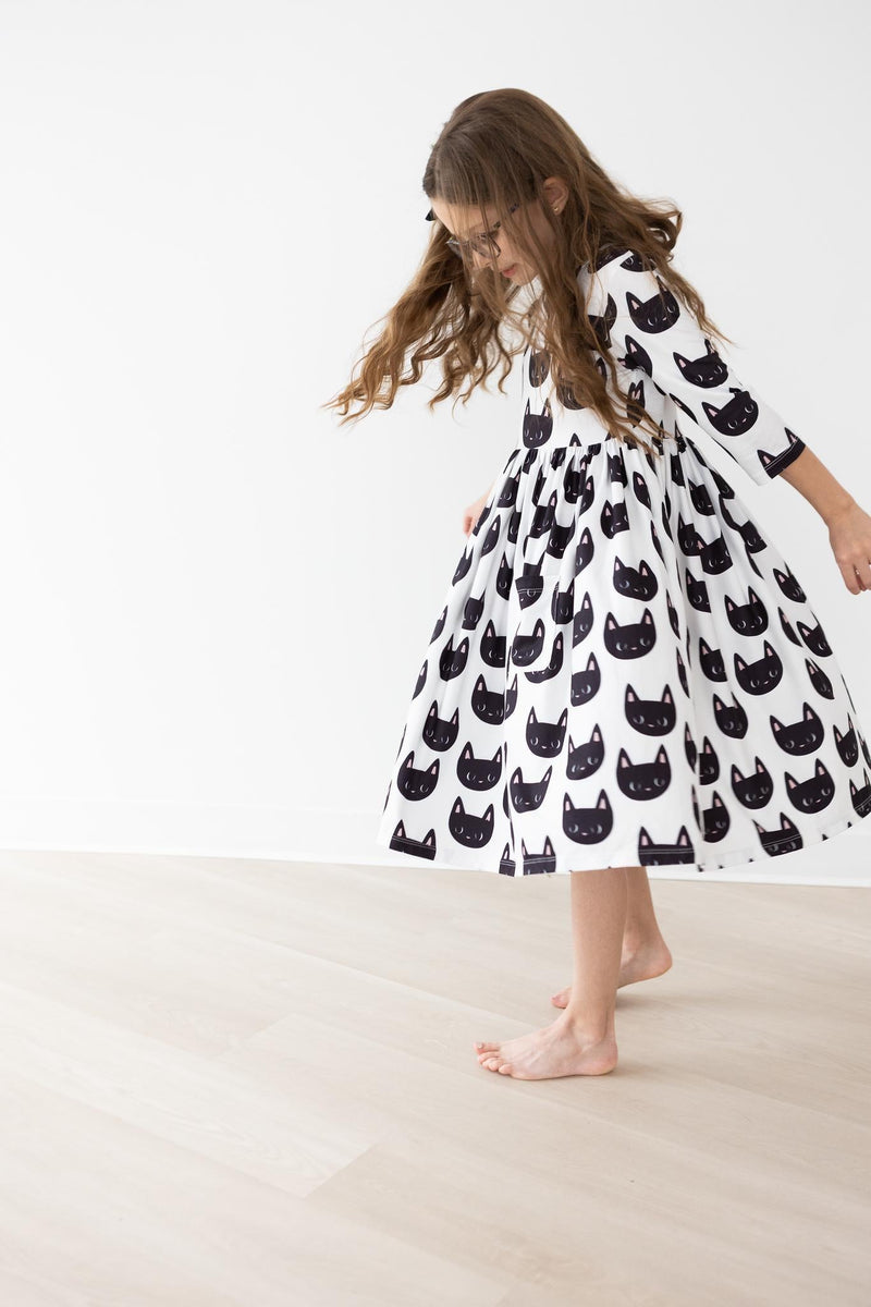 A young girl happily twirling in the Purrfect 3/4 Sleeve Pocket Twirl Dress by Mila & Rose, featuring a playful black cat print on a white background.