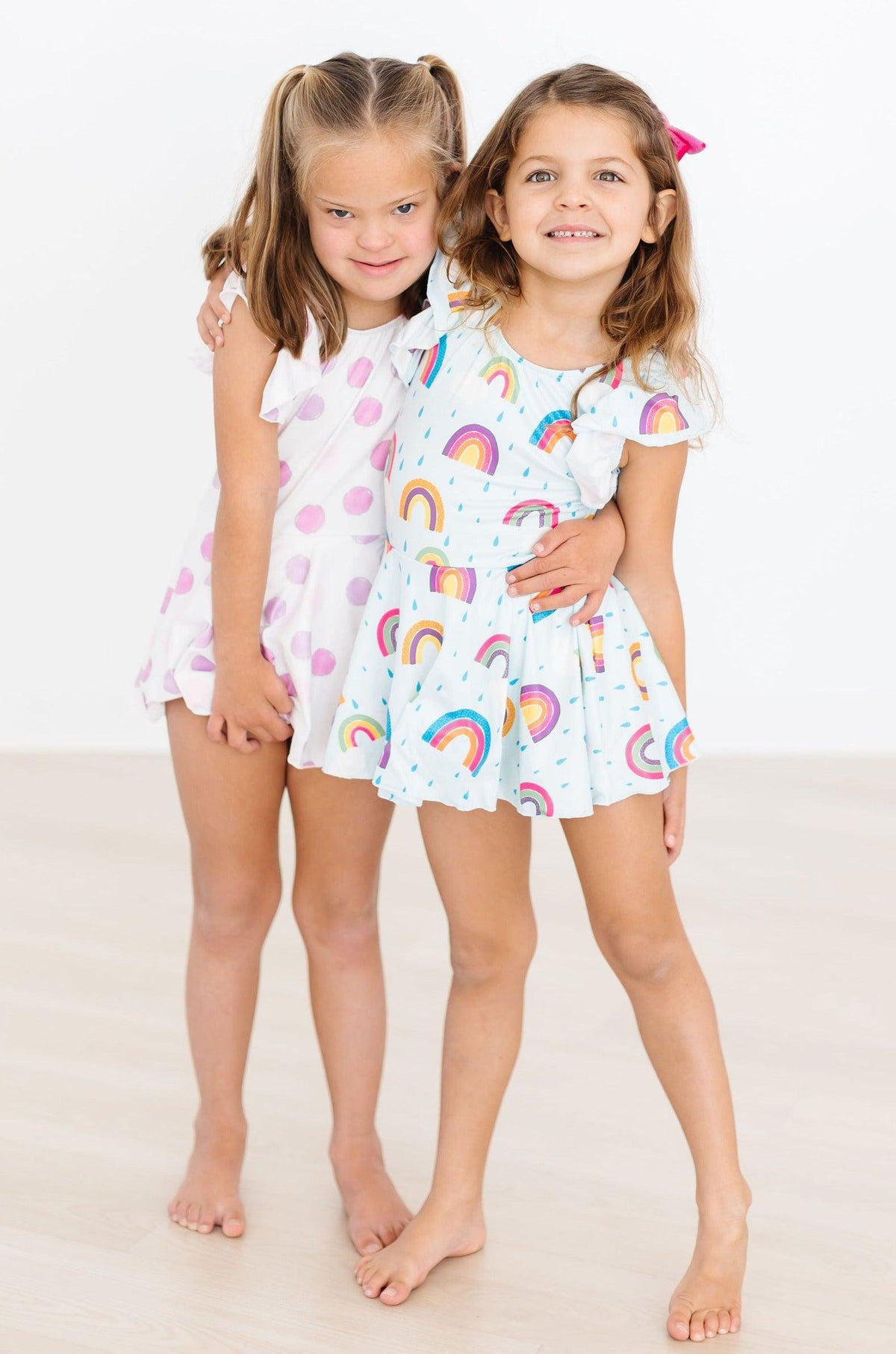 Two smiling young girls in pastel rainbow-print twirl dresses, inspired by the April Showers leotard for tiny dancers.