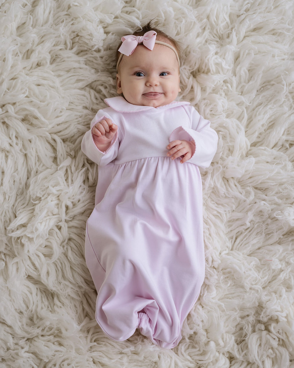 Baby wearing a pink striped classic gown with picot trim and matching bow headband on a white faux fur rug.