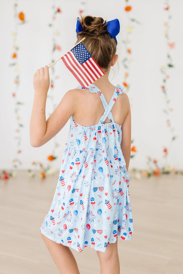 Girl wearing Baby You're a Firework Ruffle Cross Back Dress with crisscross straps and ruffle, holding an American flag.