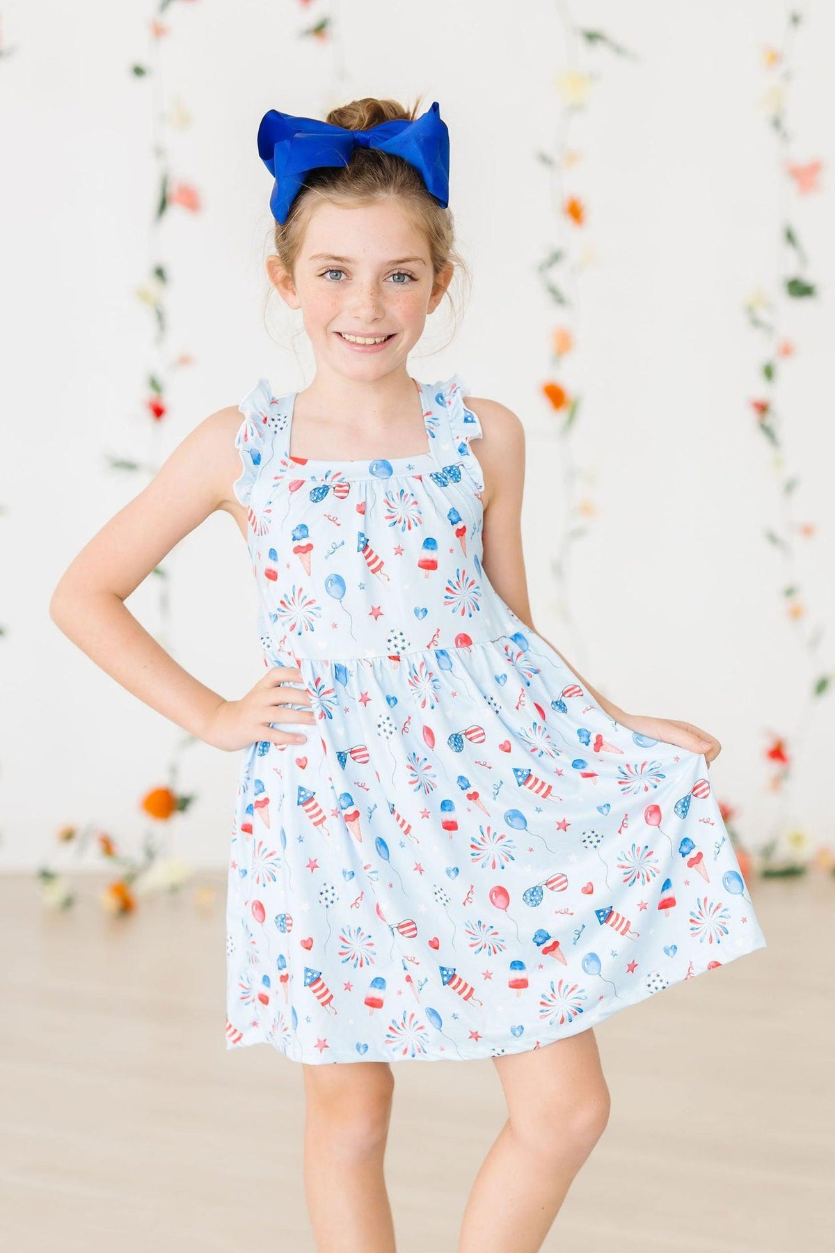Girl wearing Baby You're a Firework Ruffle Cross Back Dress with crisscross back straps and shoulder ruffle, blue bow.