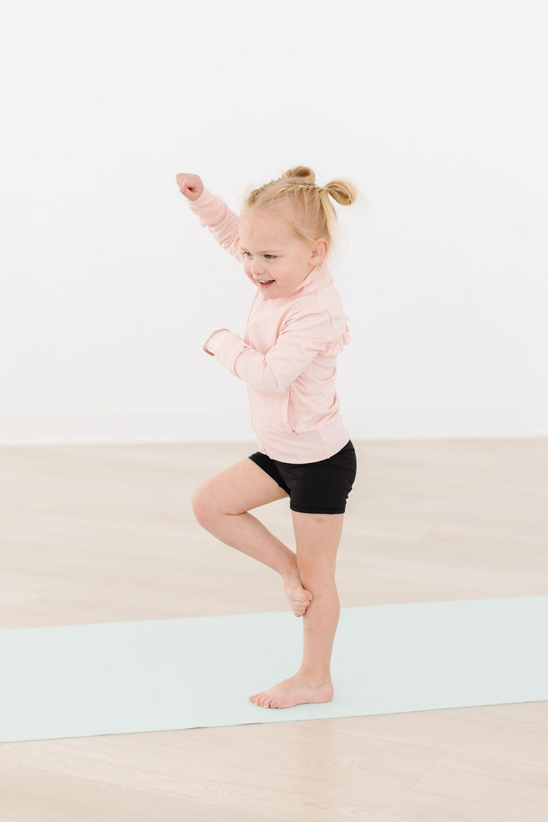 A playful girl dressed in the Zen Pink Full-Zip Ruffle Active Jacket, demonstrating an energetic pose during a yoga session, embodying fun and activity.