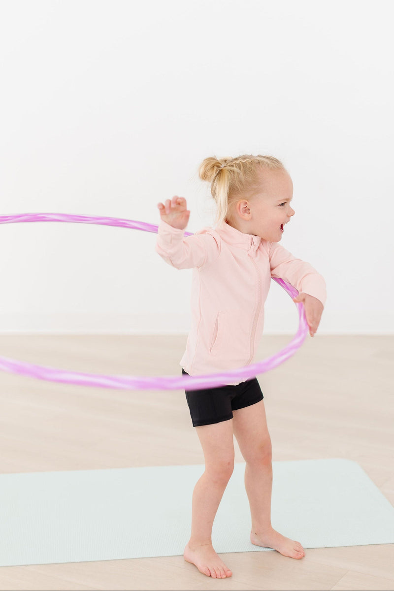 An energetic girl smiles while twirling a hula hoop, dressed in the Zen Pink Full-Zip Ruffle Active Jacket and black shorts, embodying fun and fitness.