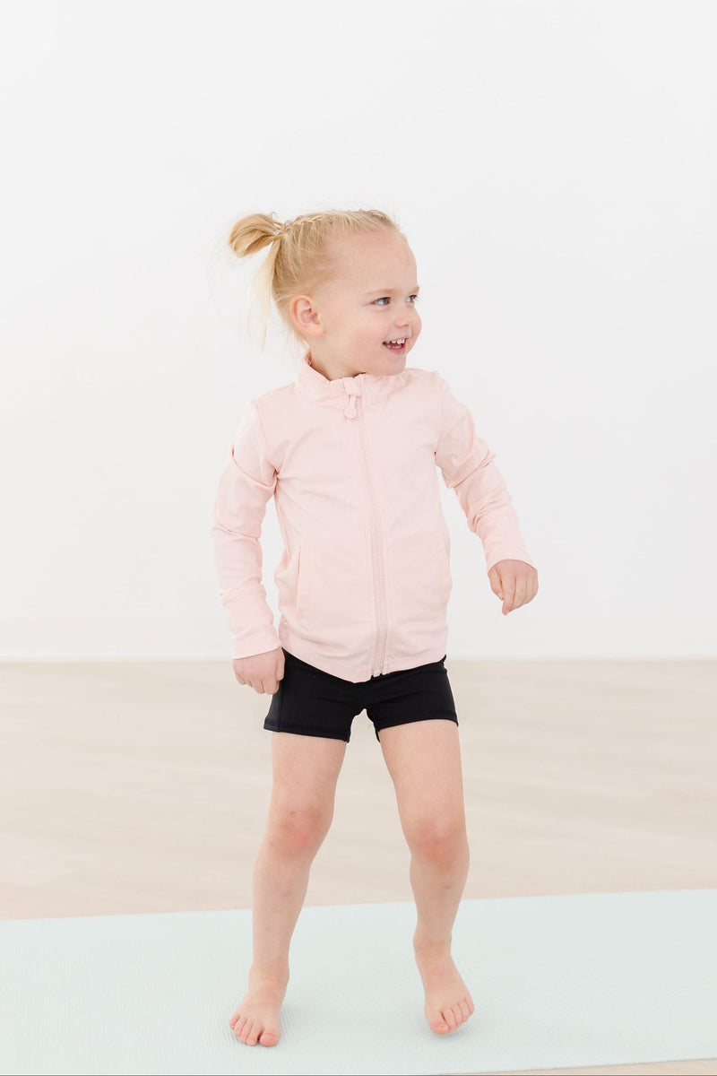 A cheerful girl in motion, wearing the Zen Pink Full-Zip Ruffle Active Jacket over black shorts, perfect for active play.