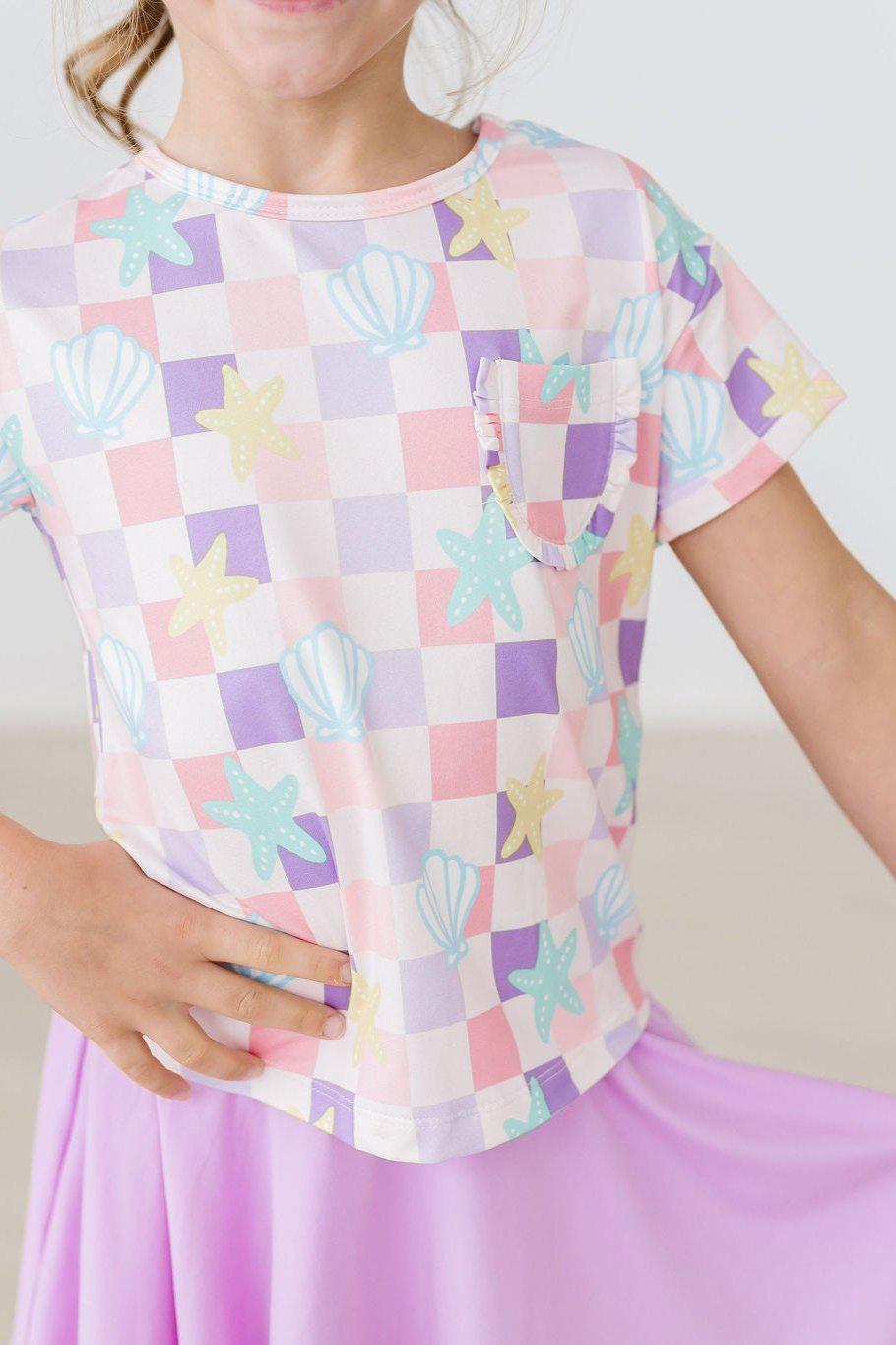 Cute girl wearing the Beach Days Ruffle Pocket Tee featuring a fun starfish and seashell print, perfect for summer playdates and adventures!