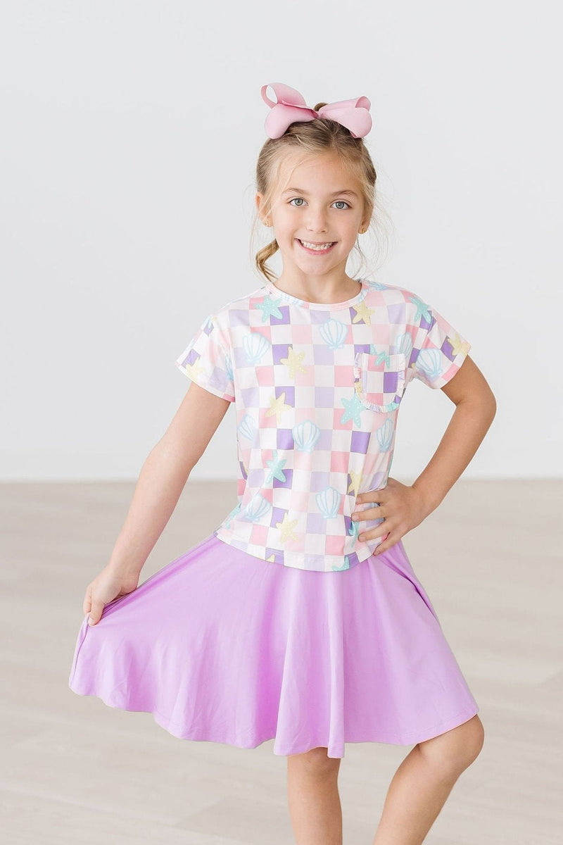 A cheerful girl modeling the Beach Days Ruffle Pocket Tee, twirling in a lovely lavender skirt, ready for playful outings.