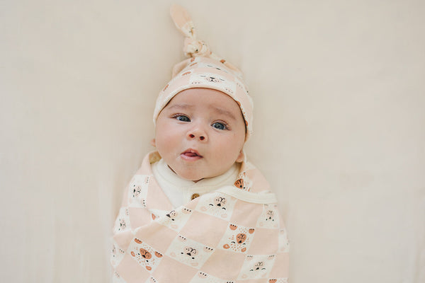 Baby wearing GOTS certified organic cotton knotted hat with checkered bear print, swaddled in matching blanket.