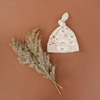 GOTS certified organic cotton baby hat with checkered bear print and knotted top, styled with dried grass on terracotta background.