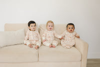 Three babies sitting on a couch wearing GOTS certified organic cotton zippy rompers with a pink checkered bear pattern.
