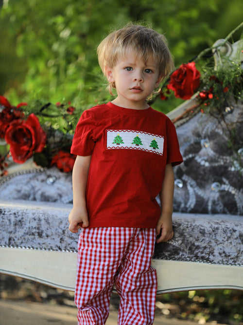 Blonde toddler boy in red tee with green tree embroidery and plaid pants standing on grey sofa