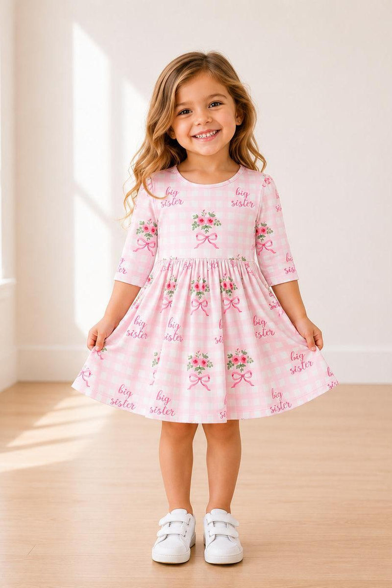 Young girl in pink gingham Big Sister Floral 3/4 sleeve twirl dress with floral bouquets and bows, smiling.