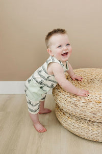 Happy baby standing by a woven pouf wearing a GOTS certified organic cotton sleeveless romper with black print and mint green pockets.