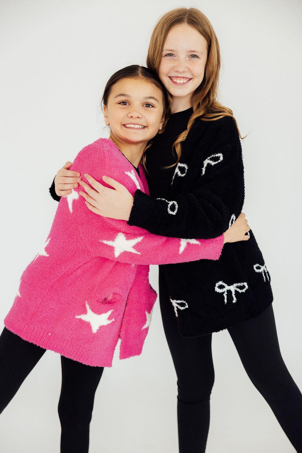 Girls hugging in fuzzy black bow and pink star cardigans. Cozy kids sweaters for comfortable styling.