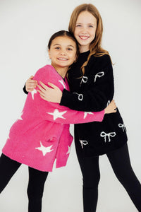 Girls hugging in fuzzy black bow and pink star cardigans. Cozy kids sweaters for comfortable styling.