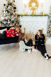 Girls model cozy black, red, and green cardigans with bow and star details near a gold and black Christmas tree.