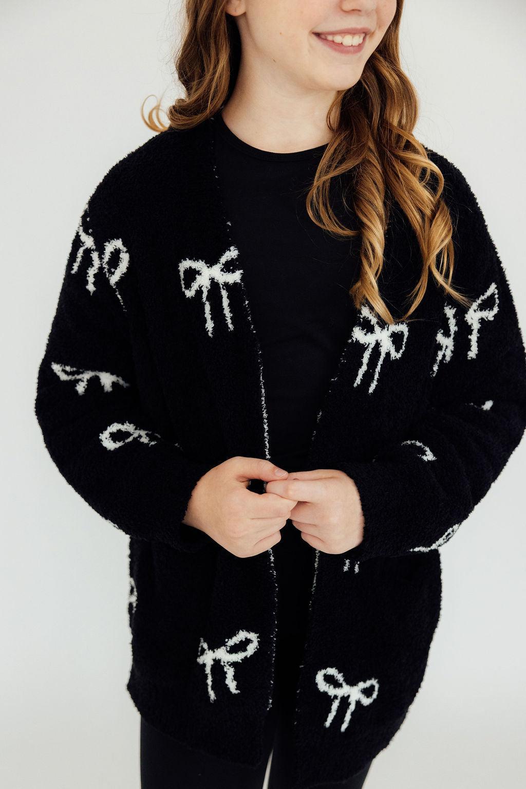 Black cozy cardigan featuring white bow patterns, styled open over a black top for a comfortable casual look.