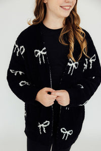 Black cozy cardigan featuring white bow patterns, styled open over a black top for a comfortable casual look.