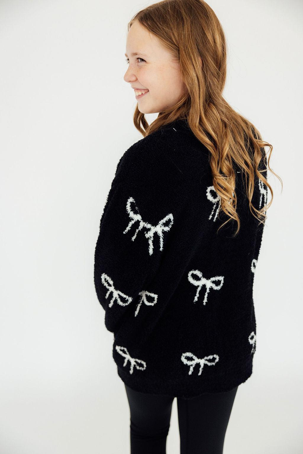 Black cozy cardigan with white bow patterns worn over black leggings for a stylish, comfortable cool-weather look.