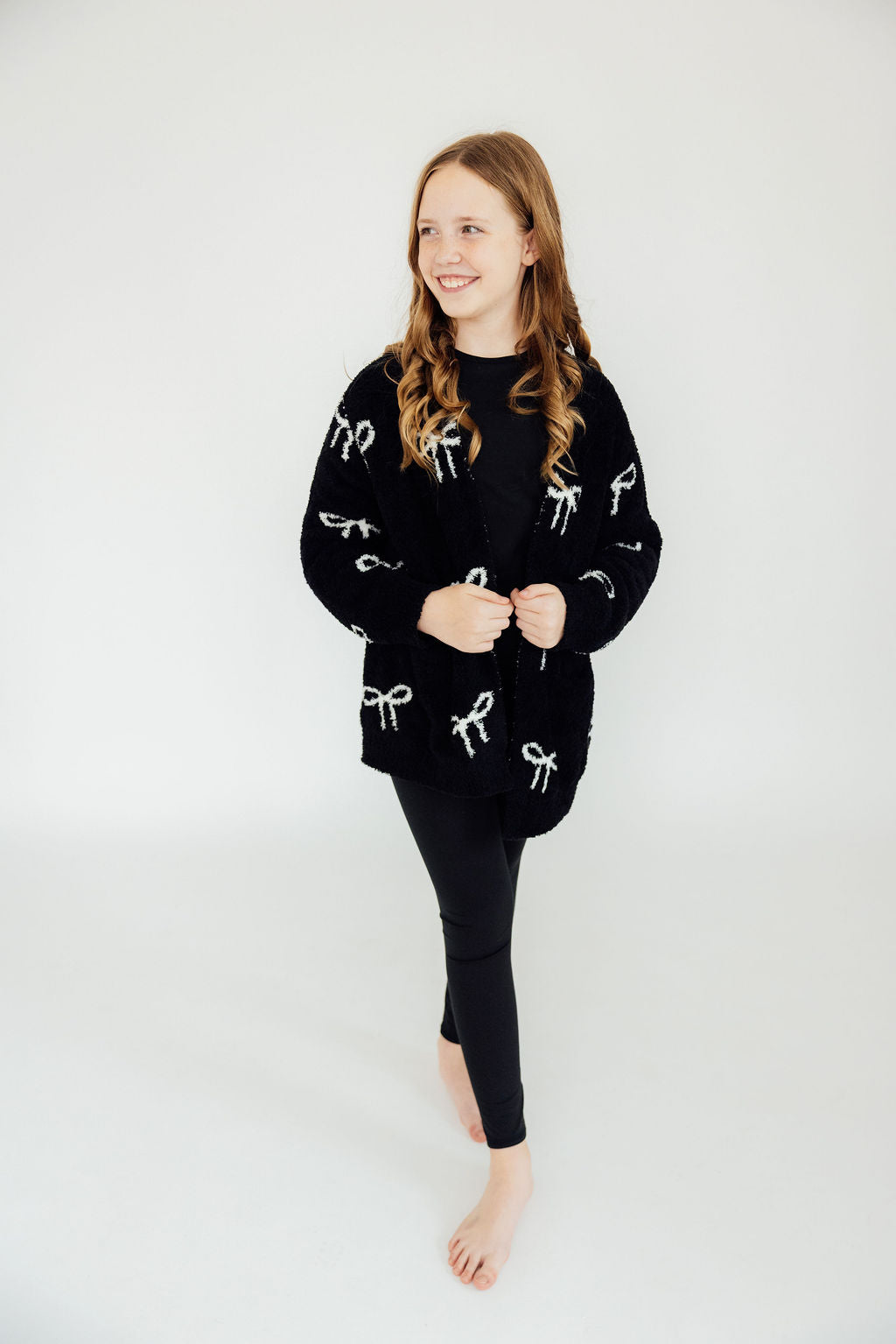 Girl wearing a black cozy cardigan with white bow patterns and black leggings. Stylish and comfortable layering piece.