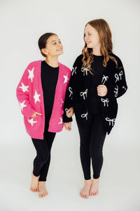 Two girls modeling cozy cardigans; one wears a black bow print style and the other a pink star print version, both paired with black leggings.